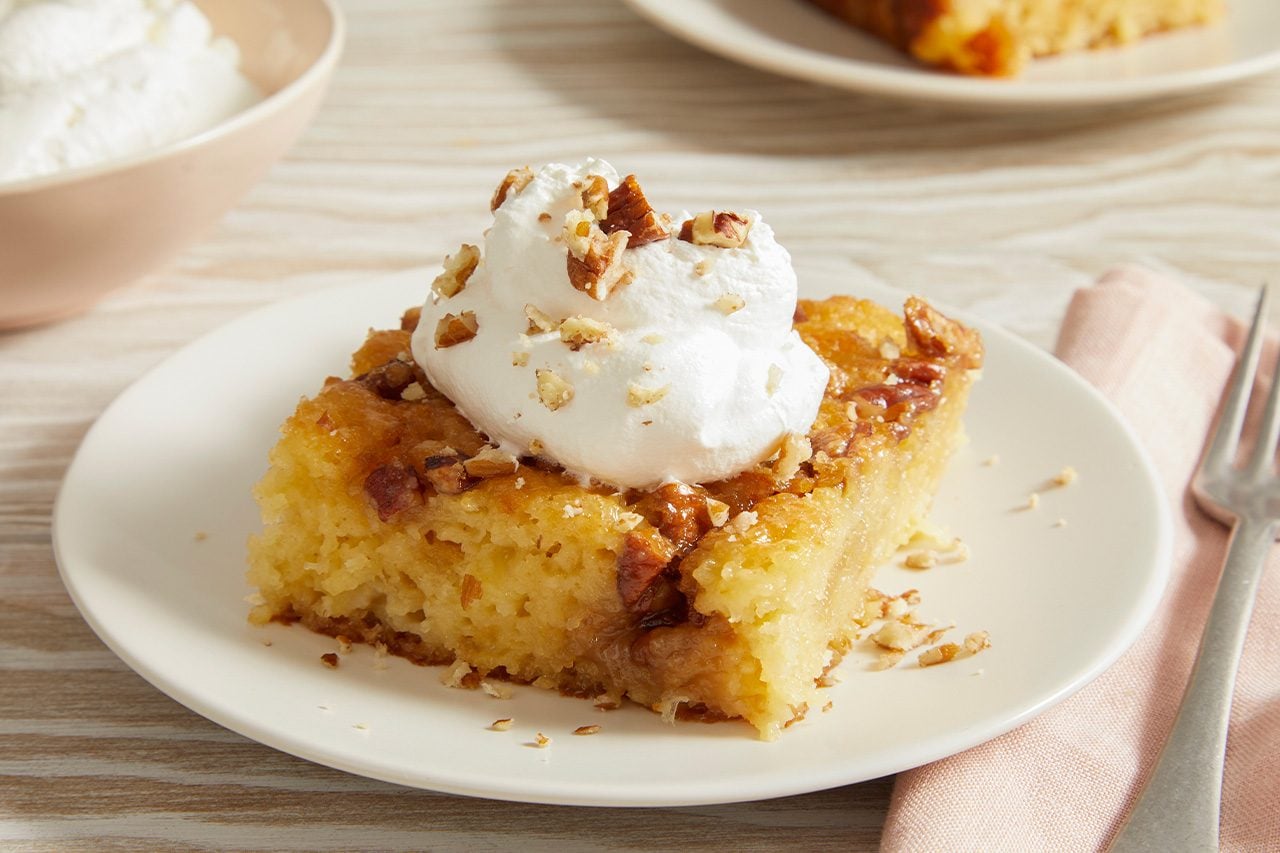 Eye-level horizontal image of a moist cake slice, drizzled with a sweet glaze and topped with a generous dollop of whipped cream and chopped nuts. The dessert is presented on a white plate, with a fork and a napkin to the side.