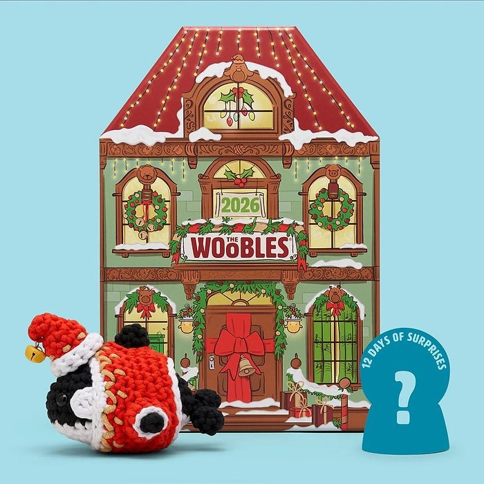 A festive house-shaped advent calendar labeled “Woobles 2026” is displayed behind a crocheted penguin in a Santa hat and a blue sign that reads “12 Days of Surprises” with a question mark.