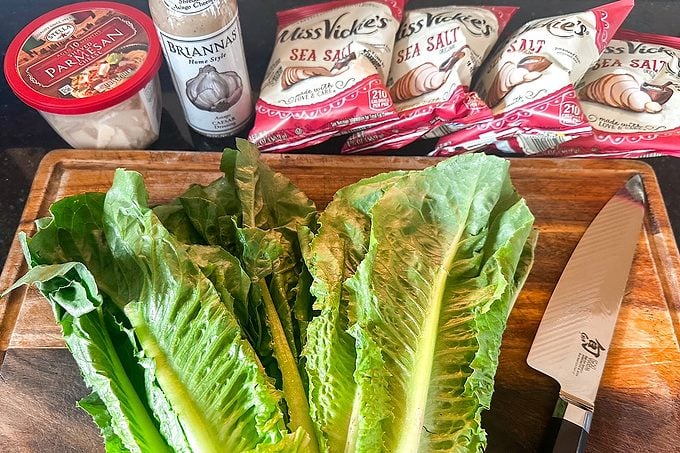 Fresh romaine lettuce on a wooden cutting board beside a chef’s knife, Parmesan cheese, dressing, and sea salt crackers, evoking meal prep.