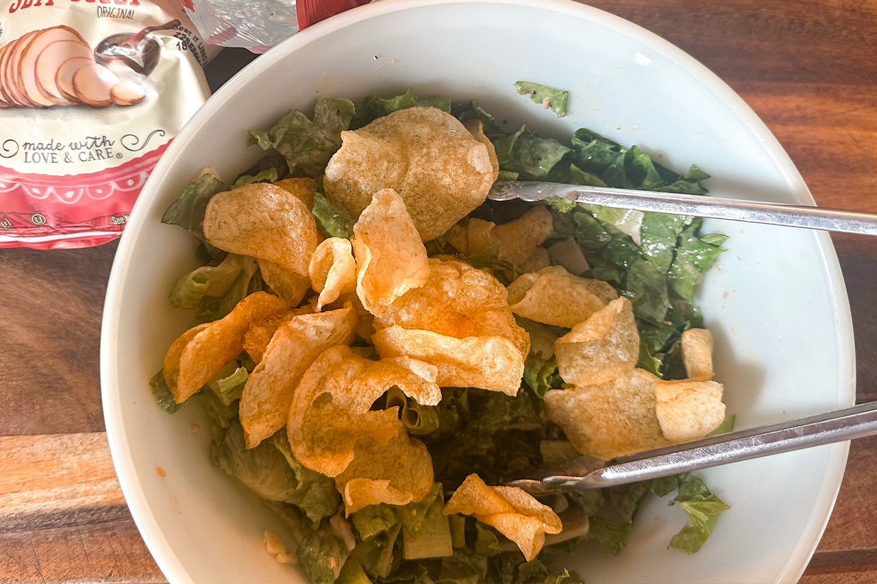 White bowl of leafy green salad topped with golden crispy chips, served with metal tongs on a wooden table; casual, appetizing meal.