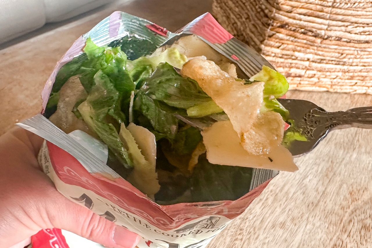 Hand holds a foil-lined paper container of fresh green salad with lettuce, pale vegetables, and crispy chips, with a fork ready to serve.