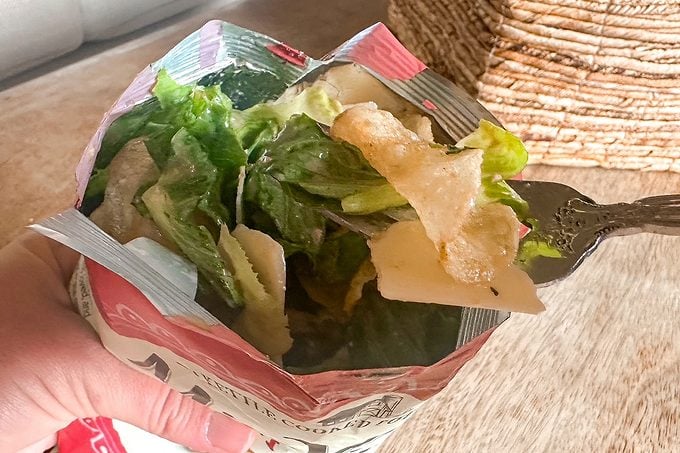 Hand holds a foil-lined paper container of fresh green salad with lettuce, pale vegetables, and crispy chips, with a fork ready to serve.