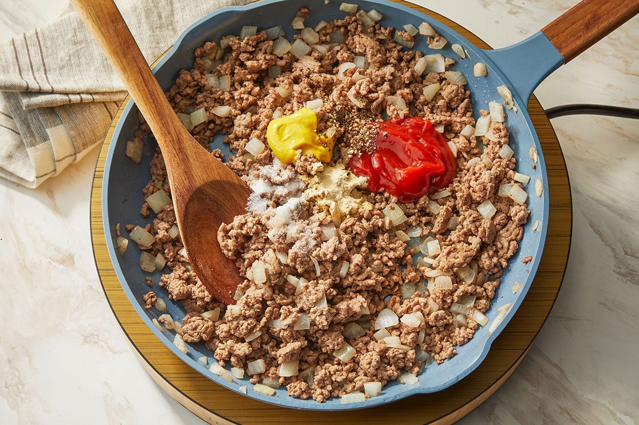 Overhead horizontal image of ground meat and chopped onions cooking in a blue pan. Ketchup, yellow mustard, salt, and various spices are added to the mixture, with a wooden spoon resting in the pan.