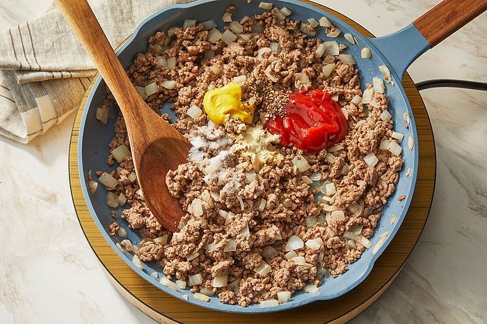 Overhead horizontal image of ground meat and chopped onions cooking in a blue pan. Ketchup, yellow mustard, salt, and various spices are added to the mixture, with a wooden spoon resting in the pan.