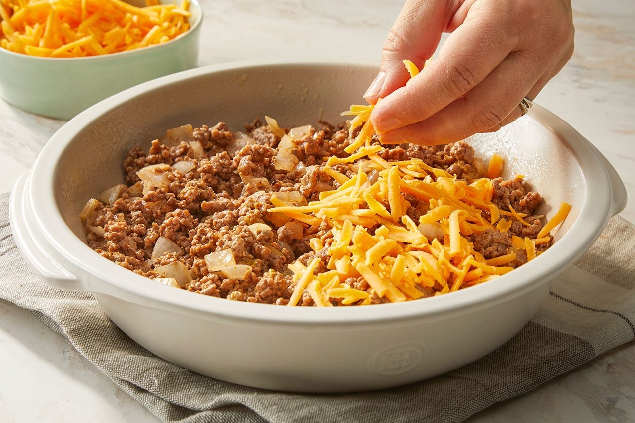 Overhead horizontal image of a hand sprinkling shredded cheese over a savory mixture of cooked ground meat and chopped white onions in a white baking dish. A bowl of additional shredded cheese is visible in the background, ready to be added to the dish.