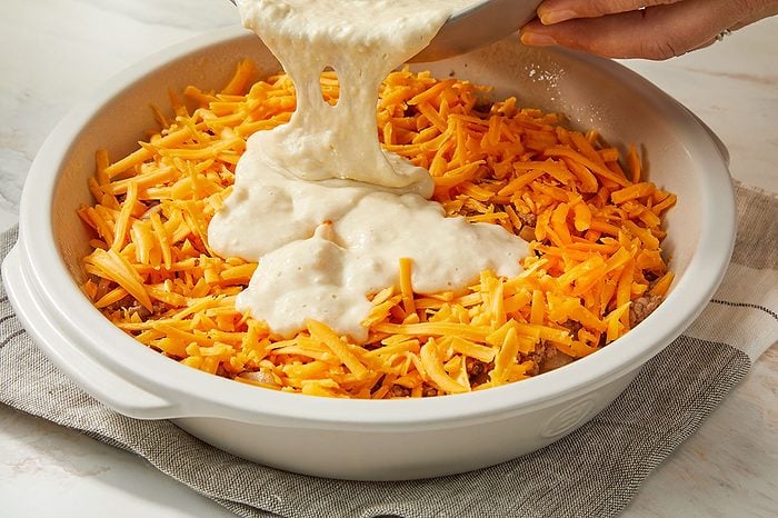 Eye Level horizontal image of a hand pouring a creamy batter from a metal bowl into a white baking dish. The dish contains a layer of shredded cheddar cheese and ground meat, with the batter being added on top.