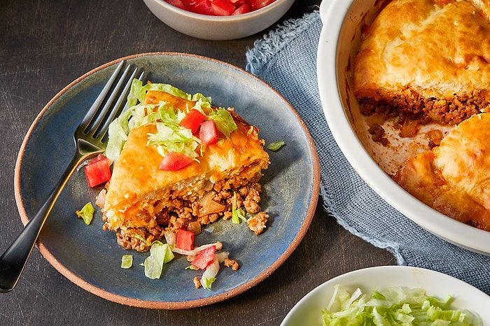 Eye level horizontal image of a slice of a savory casserole served on a blue plate with a fork. The casserole features a golden-brown topping over a ground beef and onion filling, garnished with fresh shredded lettuce and diced red tomatoes. A white baking dish with more casserole, a bowl of diced tomatoes, and a bowl of shredded lettuce are visible in the background.
