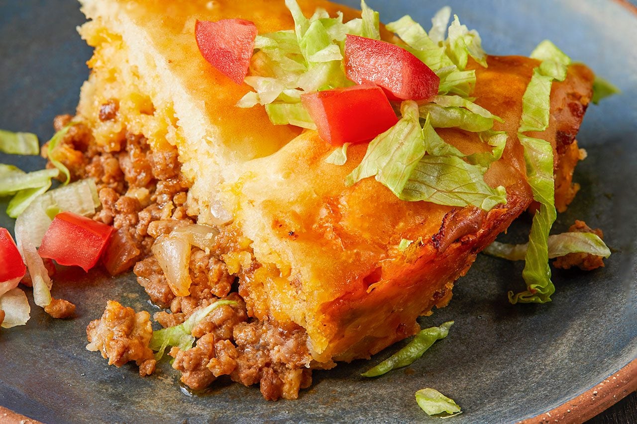 Eye Level horizontal image of a savory slice, resembling a taco pie or casserole, served on a dark blue plate. The golden-brown crust is cut open, revealing a hearty filling of seasoned ground meat and chopped onions. The dish is garnished generously with fresh shredded lettuce and diced red tomatoes.