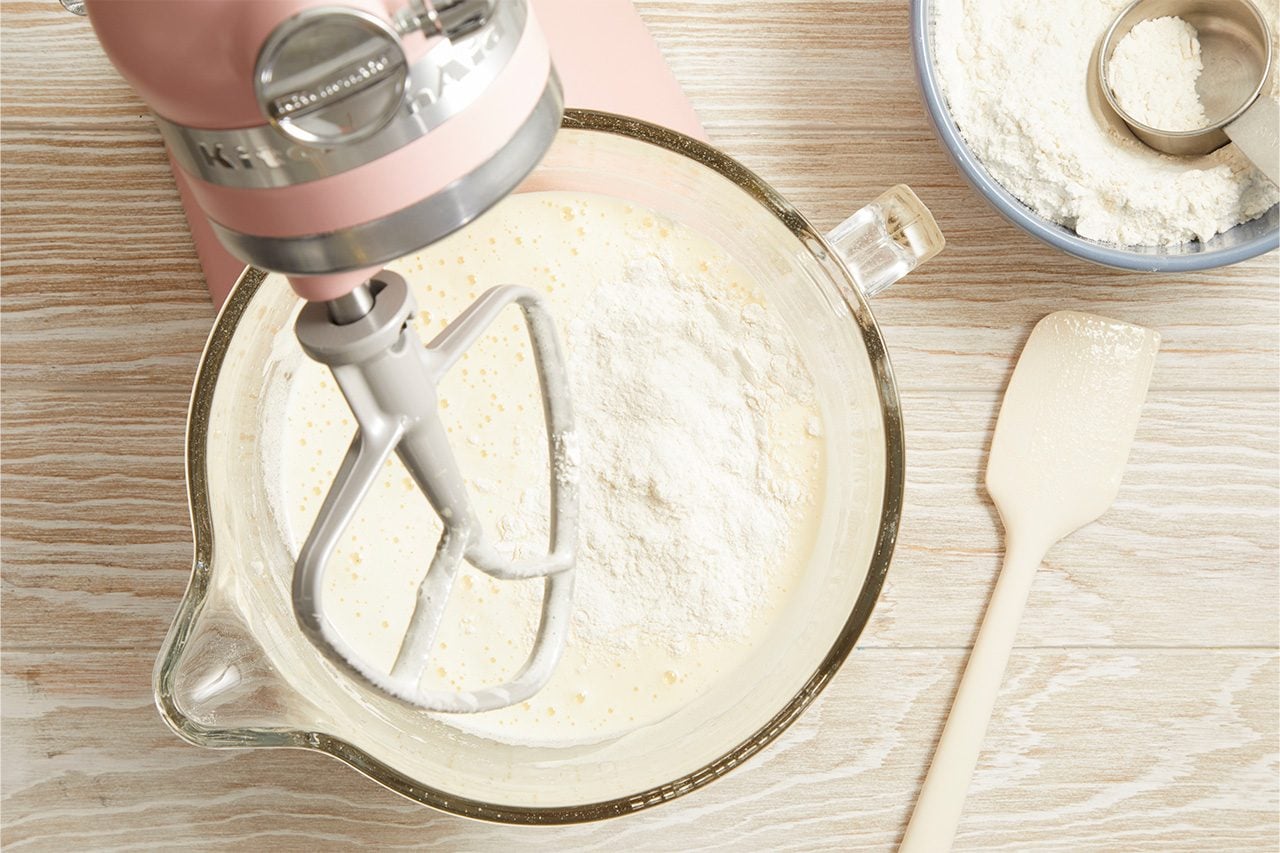 Overhead horizontal image of a pink KitchenAid stand mixer combining dry ingredients into a wet batter in a clear glass mixing bowl. A pile of white flour is being incorporated into the creamy, bubbly mixture, with additional flour visible in a blue bowl and a measuring cup to the right, next to a cream-colored spatula.