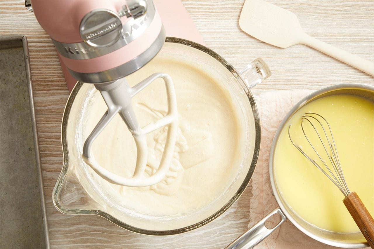Overhead horizontal image of a pink stand mixer combining a light-colored batter in a clear glass bowl. Next to it, a metal saucepan holds a yellow liquid with a whisk resting inside, and a baking sheet and silicone spatula are also visible on the light wood surface.