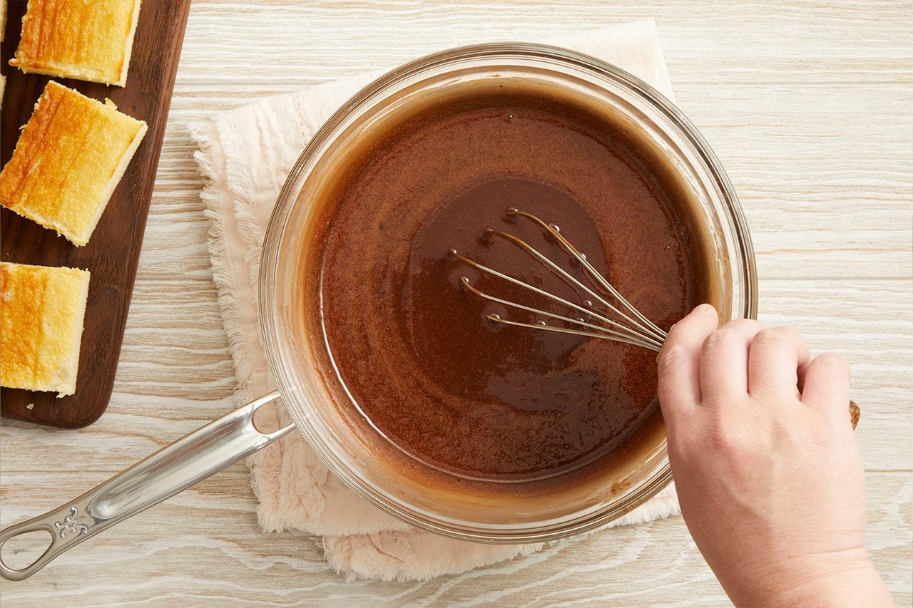Overhead horizontal image of a hand whisking a dark brown liquid mixture in a clear glass saucepan. To the left, several golden-brown baked bars are arranged on a dark wooden cutting board. The mixture is likely a chocolate sauce, being prepared alongside the finished baked goods.