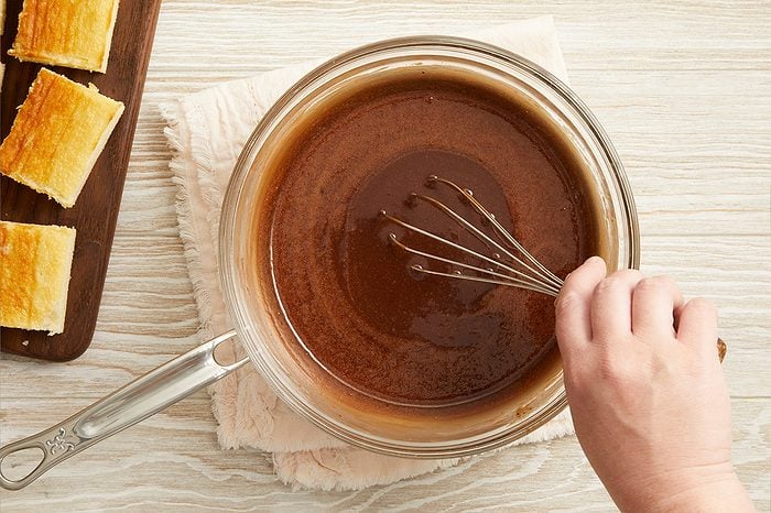 Overhead horizontal image of a hand whisking a dark brown liquid mixture in a clear glass saucepan. To the left, several golden-brown baked bars are arranged on a dark wooden cutting board. The mixture is likely a chocolate sauce, being prepared alongside the finished baked goods.