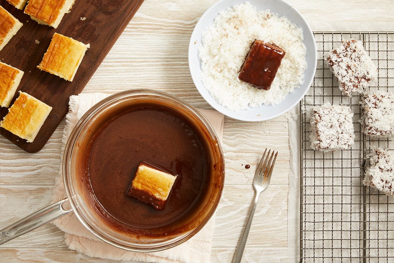 Overhead horizontal image of square cake pieces being prepared. One cake piece is partially dipped into a clear bowl of chocolate sauce, while another chocolate-coated piece rests in a bowl of shredded coconut. Several finished desserts, coated in chocolate and coconut, are arranged on a wire rack next to a metal fork.