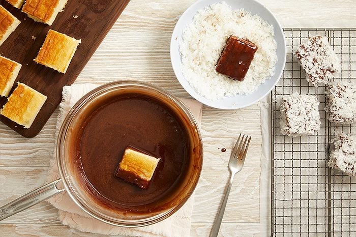 Overhead horizontal image of square cake pieces being prepared. One cake piece is partially dipped into a clear bowl of chocolate sauce, while another chocolate-coated piece rests in a bowl of shredded coconut. Several finished desserts, coated in chocolate and coconut, are arranged on a wire rack next to a metal fork.