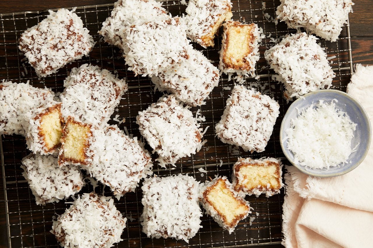 Overhead horizontal image of numerous coconut-covered square cakes arranged on a metal cooling rack. Several cakes are cut in half, revealing a light yellow sponge cake interior beneath a dark coating and a thick layer of shredded coconut. A small blue bowl filled with shredded coconut and a pink cloth are also visible on the dark wooden surface.