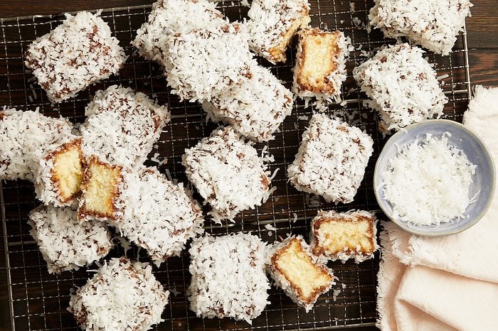 Overhead horizontal image of numerous coconut-covered square cakes arranged on a metal cooling rack. Several cakes are cut in half, revealing a light yellow sponge cake interior beneath a dark coating and a thick layer of shredded coconut. A small blue bowl filled with shredded coconut and a pink cloth are also visible on the dark wooden surface.