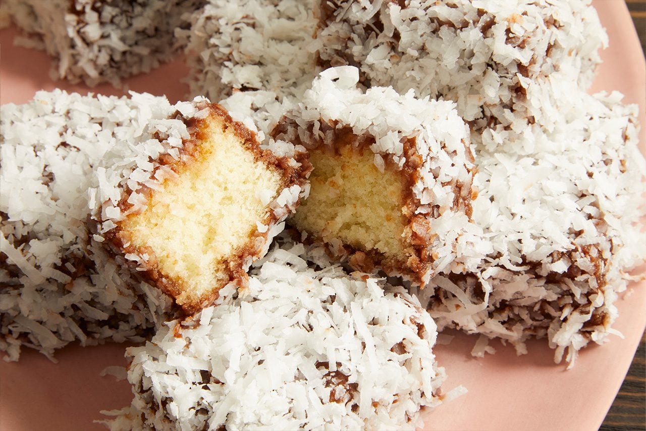 Eye Level horizontal image of multiple square-shaped cake pieces heavily coated in shredded coconut and a dark brown layer, resting on a pink plate. One piece is broken open, revealing its light yellow cake interior.