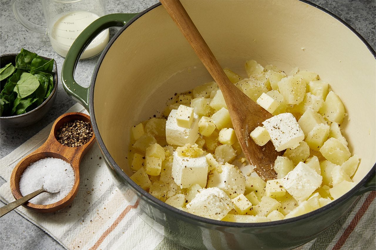 Overhead horizontal image of cubed cooked potatoes, butter, and cream cheese in a green Dutch oven, seasoned with pepper. A wooden spoon rests in the pot, with a bowl of fresh spinach, a measuring cup of milk, and a salt and pepper cellar visible on the counter.