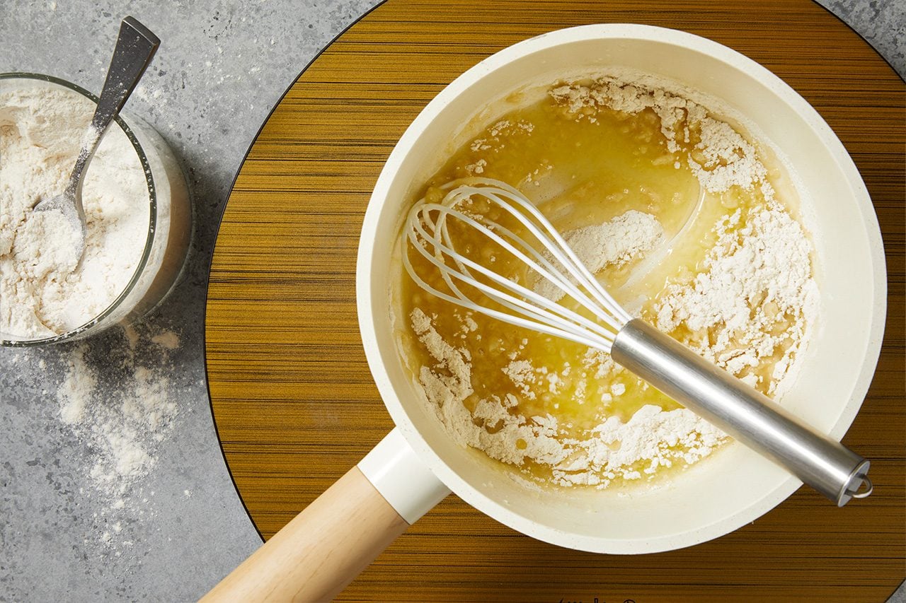 Overhead horizontal image of a white saucepan containing melted butter and flour, with a whisk resting inside. To the left, a clear glass bowl holds additional flour with a spoon.