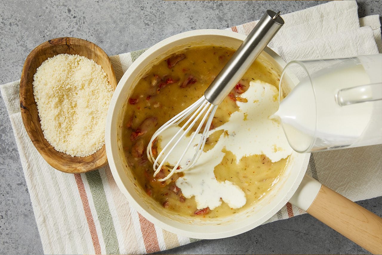 Overhead horizontal image of milk or cream being poured from a glass measuring cup into a white pan containing a sauce with sun-dried tomatoes and herbs. A white whisk rests in the pan, and a wooden bowl of grated Parmesan cheese sits nearby on a striped tea towel.