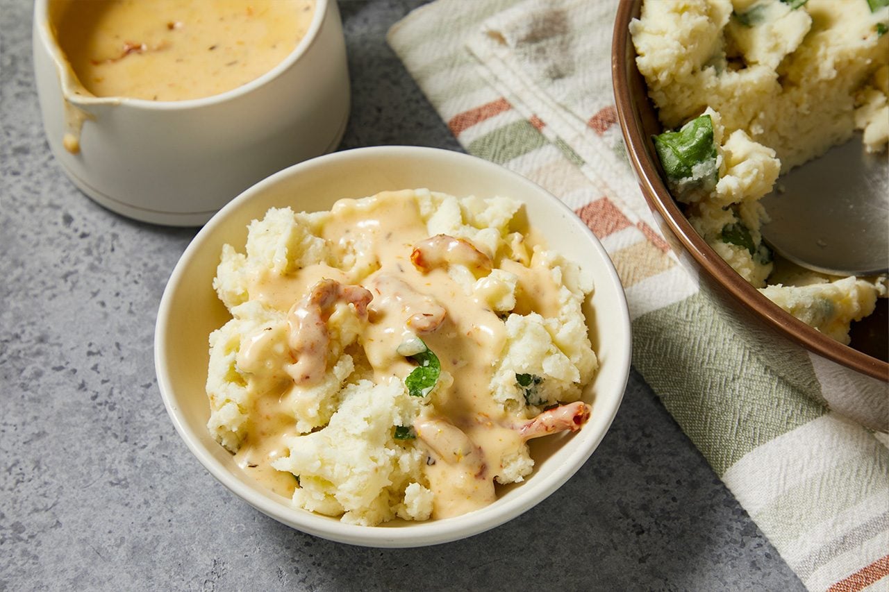 Eye Level horizontal image of a bowl of mashed potatoes topped with a creamy sauce. The sauce contains pieces of what appear to be sun-dried tomatoes. In the background, a gravy boat holds more sauce, and a serving dish with mashed potatoes mixed with green herbs is also visible.