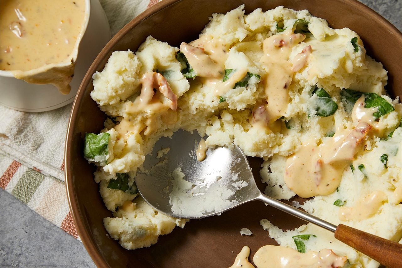 Overhead horizontal image of a brown serving bowl filled with creamy mashed potatoes, mixed with green spinach and reddish pieces. A rich, light-colored sauce is drizzled generously over the potatoes, with a metal serving spoon resting in the bowl after taking a scoop. A gravy boat containing more sauce is visible in the background.
