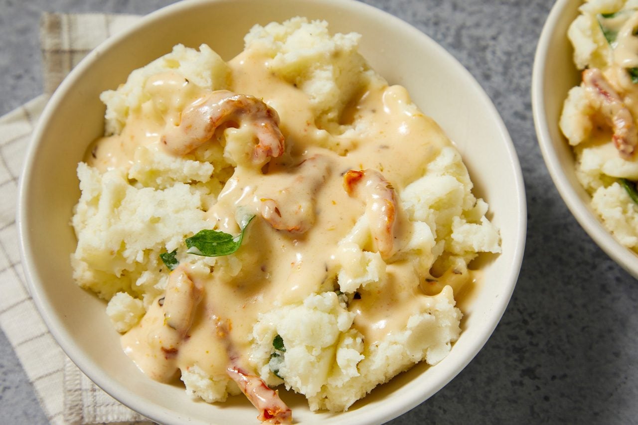 Overhead horizontal image of a bowl of mashed potatoes topped with a creamy sauce, shrimp, and green herbs. The comforting dish is presented in a light-colored bowl, resting on a gray surface with a checkered cloth in the background.
