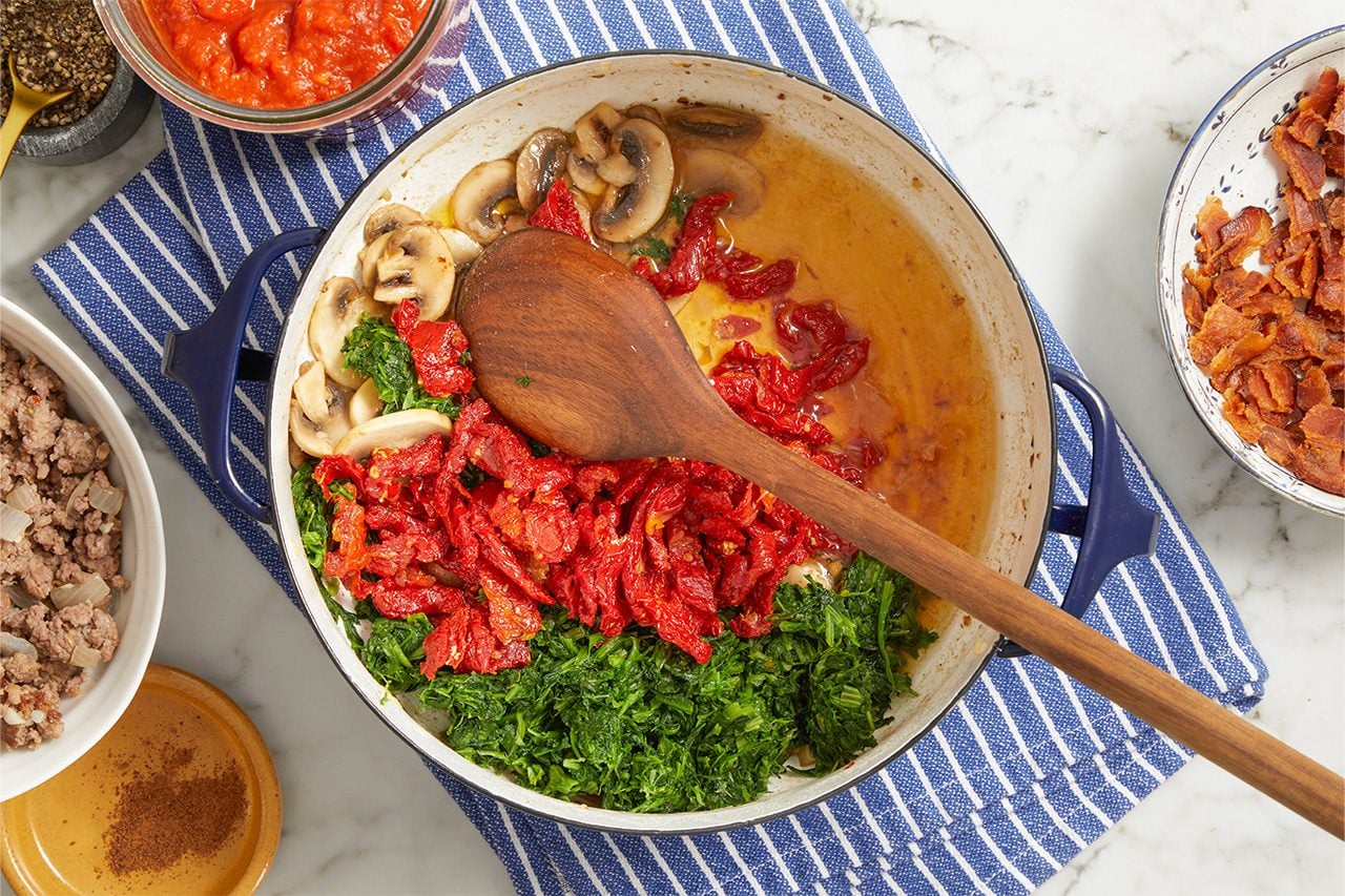 Overhead horizontal image of various ingredients arranged around and inside a blue-handled pan on a striped towel. Sliced mushrooms, chopped greens, and sun-dried tomatoes are gathered within the pan, with a wooden spoon resting on top. Bowls of cooked ground meat with onions, bacon bits, and tomato sauce are positioned around the pan, alongside ground pepper and a small dish of ground nutmeg.