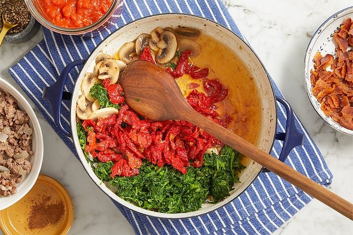 Overhead horizontal image of various ingredients arranged around and inside a blue-handled pan on a striped towel. Sliced mushrooms, chopped greens, and sun-dried tomatoes are gathered within the pan, with a wooden spoon resting on top. Bowls of cooked ground meat with onions, bacon bits, and tomato sauce are positioned around the pan, alongside ground pepper and a small dish of ground nutmeg.