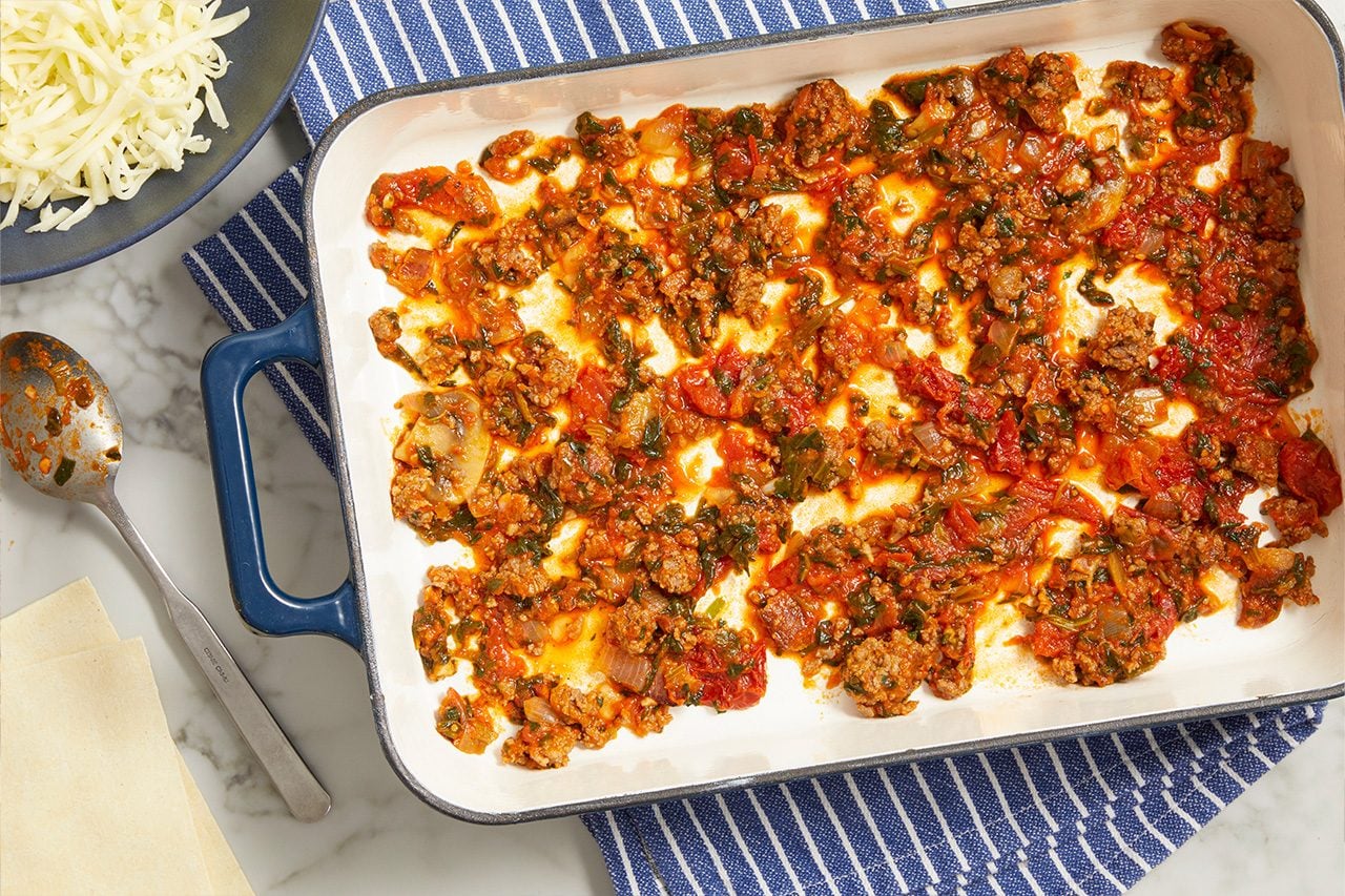 Overhead horizontal image of a white baking dish containing a layer of meat sauce. The sauce, made with ground meat, diced tomatoes, onions, and greens, covers the bottom of the dish. To the left, shredded cheese is on a dark plate, and uncooked lasagna noodles sit on a marble surface next to a sauce-covered spoon.