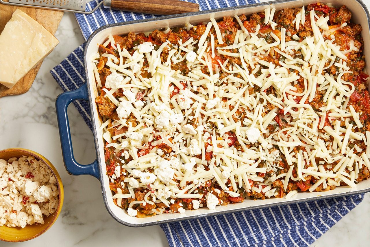 Overhead horizontal image of a baking dish filled with a savory ground meat and vegetable mixture, topped generously with shredded cheese and crumbled feta. To the left, a block of Parmesan cheese, a grater, and a small bowl of crumbled feta cheese are visible on a marble surface.