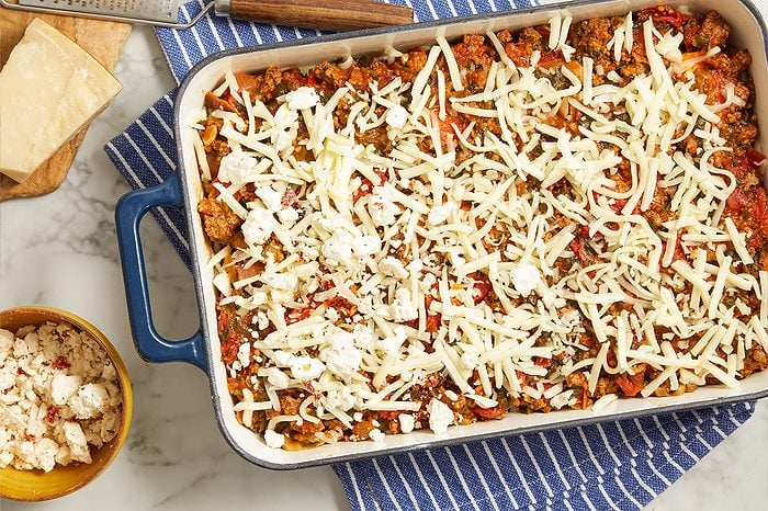 Overhead horizontal image of a baking dish filled with a savory ground meat and vegetable mixture, topped generously with shredded cheese and crumbled feta. To the left, a block of Parmesan cheese, a grater, and a small bowl of crumbled feta cheese are visible on a marble surface.