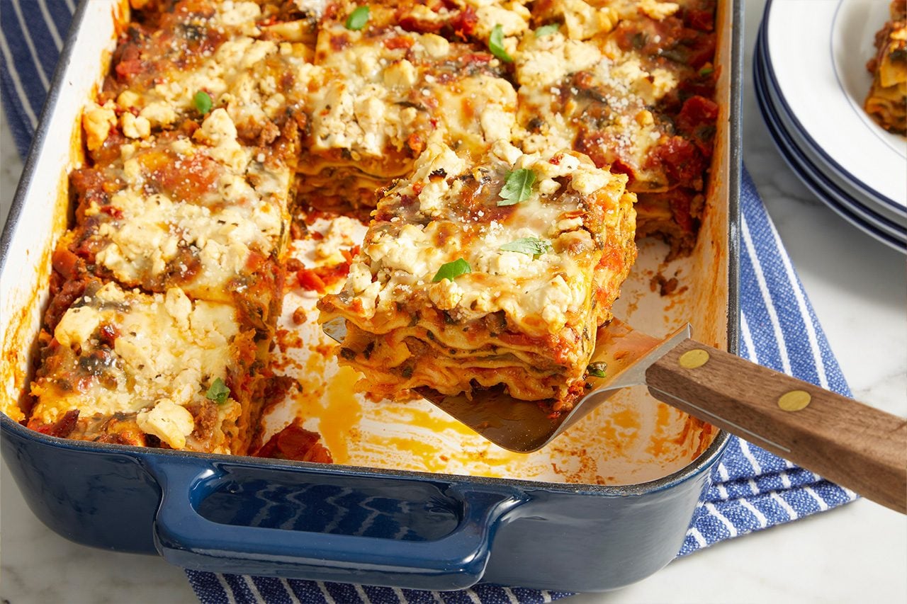 Eye level horizontal image of a serving of lasagna being lifted out of a blue baking dish with a spatula. The multi-layered lasagna features ricotta cheese, tomato sauce, and ground meat, garnished with fresh herbs and grated cheese. The casserole dish rests on a striped kitchen towel, with a stack of plates visible in the background.