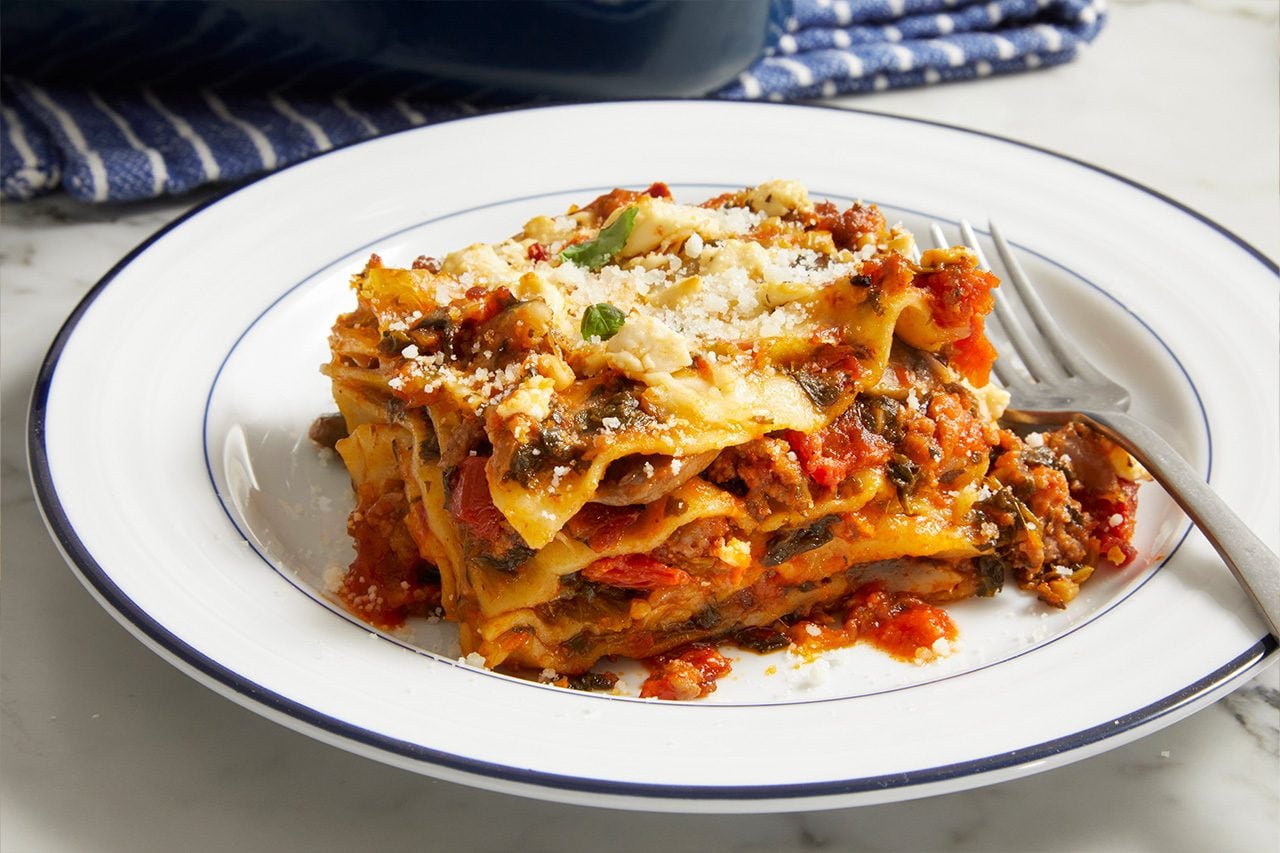 Eye level horizontal image of a generous slice of lasagna served on a white plate with a blue rim. The lasagna features multiple layers of pasta, a rich tomato-based sauce, spinach, and meat, topped with crumbled white cheese, grated Parmesan, and a fresh herb leaf. A fork rests beside the dish.