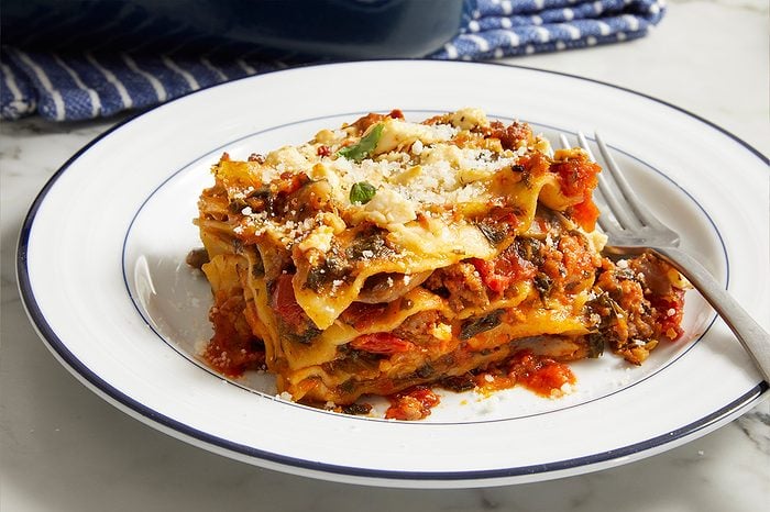 Eye level horizontal image of a generous slice of lasagna served on a white plate with a blue rim. The lasagna features multiple layers of pasta, a rich tomato-based sauce, spinach, and meat, topped with crumbled white cheese, grated Parmesan, and a fresh herb leaf. A fork rests beside the dish.