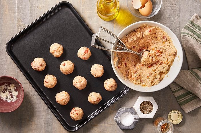 Several uncooked meatballs are arranged on a black baking sheet, while a disher rests in a bowl of meatball mixture next to olive oil, eggshells, salt, pepper, red pepper flakes, and grated cheese.