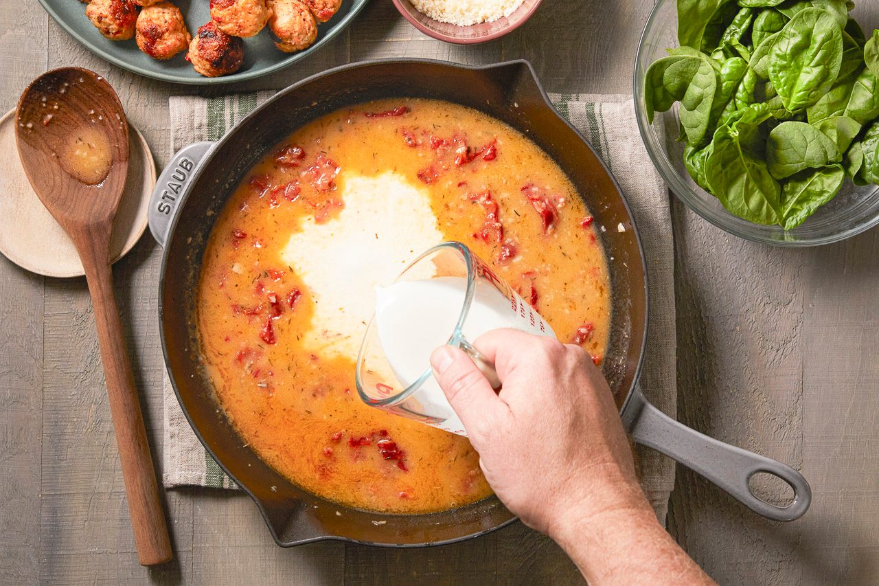 Overhead horizontal image of a hand pouring cream from a glass measuring cup into a dark cast iron pan filled with a simmering red sauce containing diced tomatoes. In the background, there are cooked meatballs, a bowl of fresh spinach, and grated Parmesan cheese.