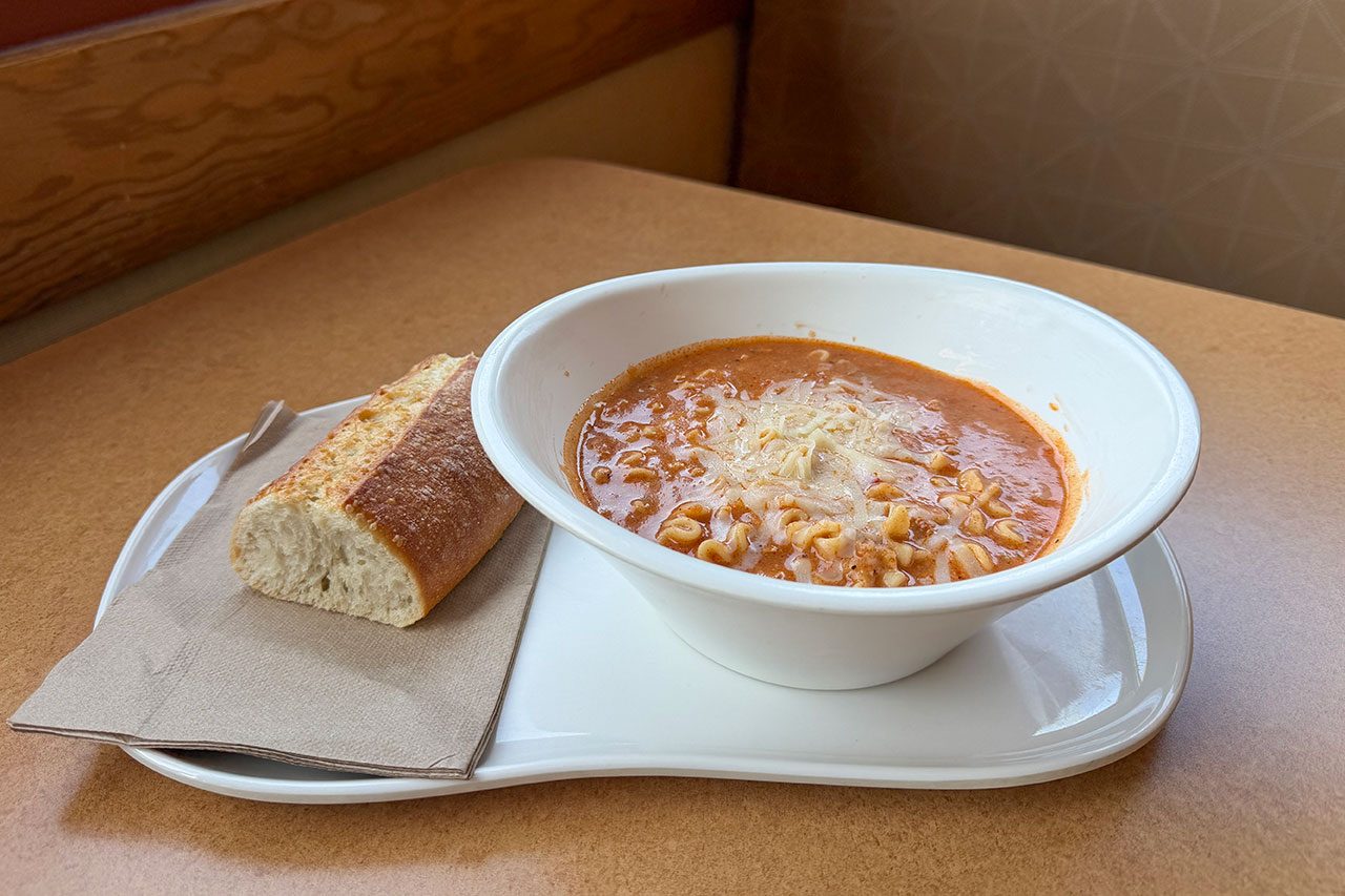 A white bowl of tomato-based soup with grated cheese on top is served on a plate with a slice of bread and a napkin, set on a light brown table.