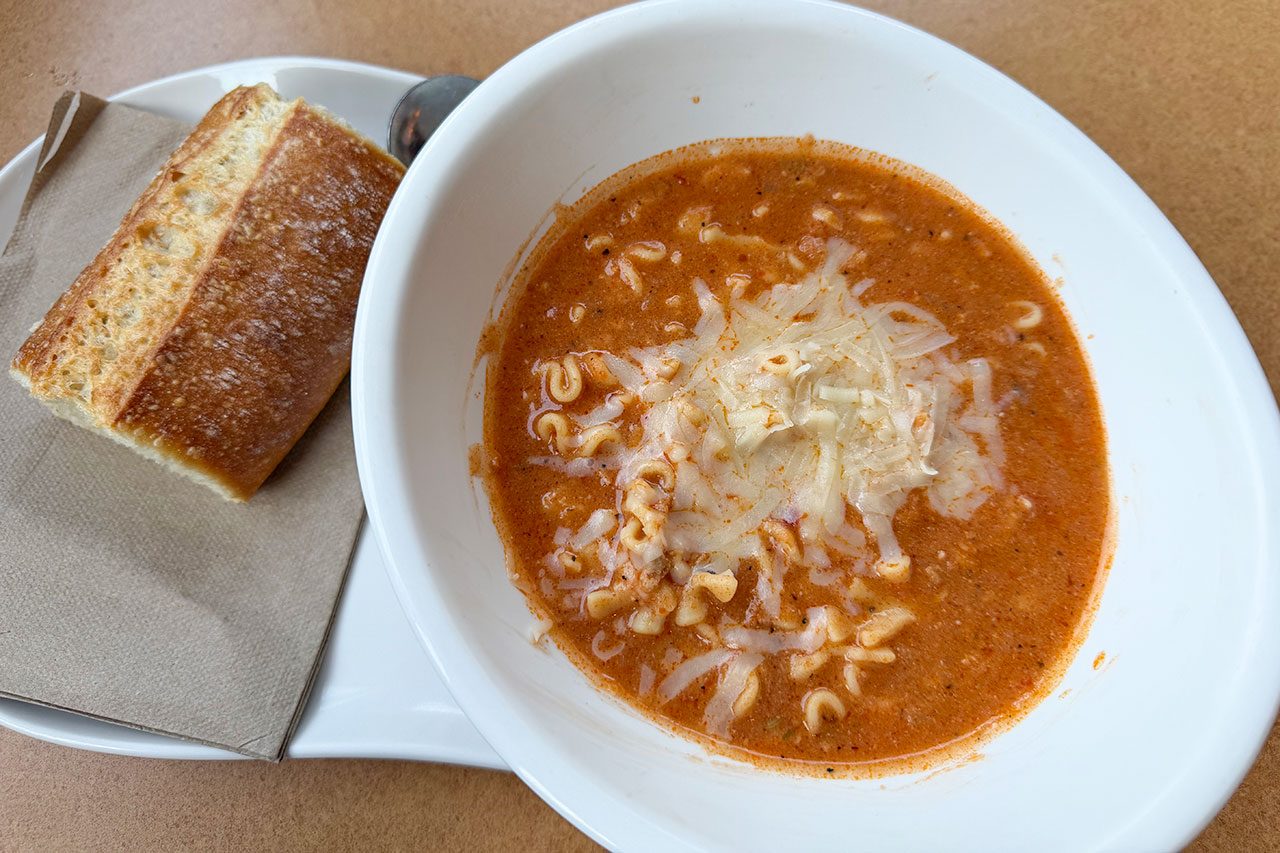 A white bowl of tomato-based soup topped with shredded cheese, served with a piece of bread on a napkin and a spoon on a white plate.