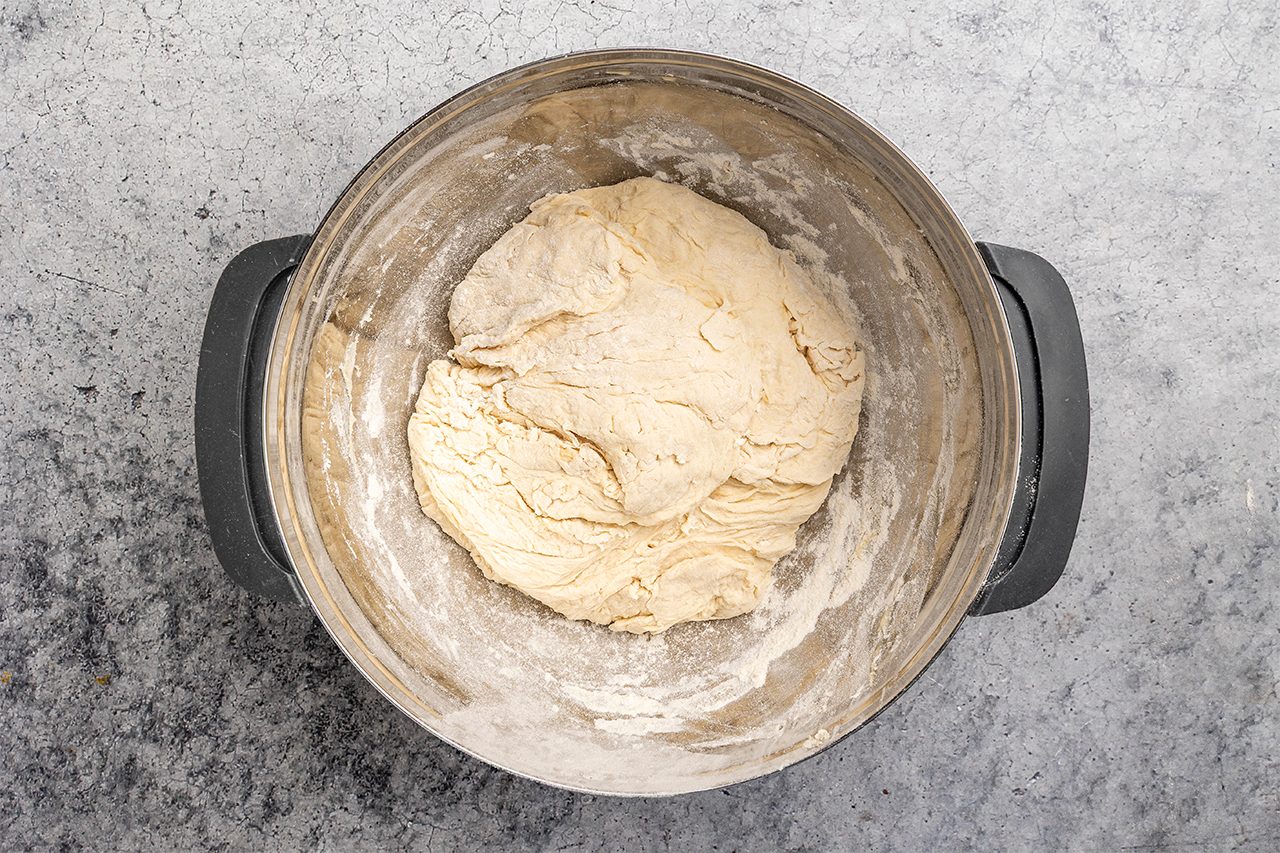 overhead shot of A stainless steel mixing bowl contains a ball of raw dough on a lightly floured surface, ready for kneading, with some flour dusting the sides of the bowl