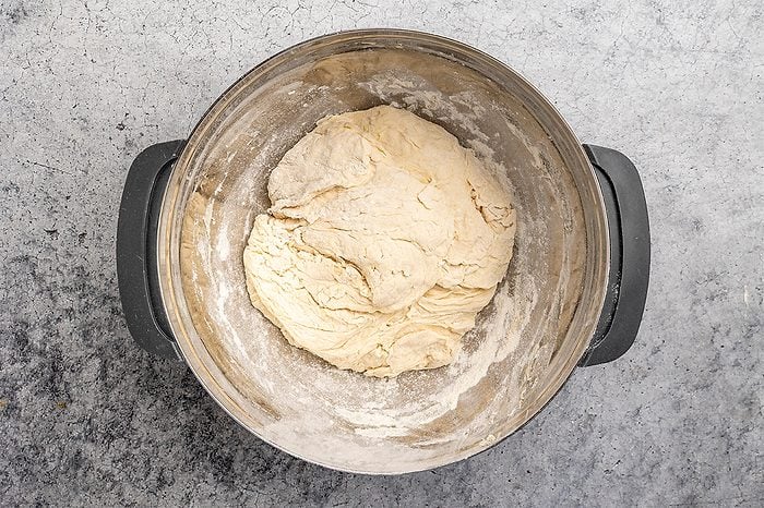 overhead shot of A stainless steel mixing bowl contains a ball of raw dough on a lightly floured surface, ready for kneading, with some flour dusting the sides of the bowl