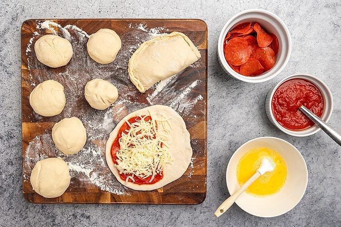 overhead shot of Round dough balls, an open calzone with sauce and cheese, and a closed calzone rest on a floured wooden board; Bowls nearby hold pepperoni, tomato sauce with a spoon, and a bowl of beaten egg with a brush