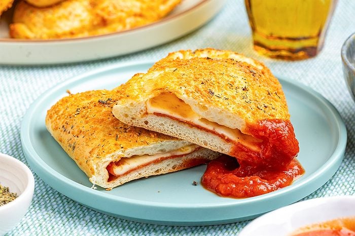 A cheesy calzone cut in half on a blue plate, with melted cheese and sauce inside; One half is dipped in tomato sauce, and there are herbs sprinkled on the crust; A glass of drink and other food are in the background
