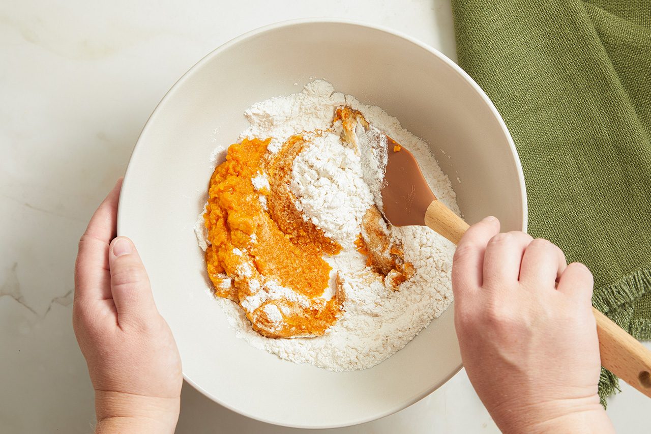 Overhead horizontal image of hands mixing ingredients in a bowl. A person's hands are holding a beige mixing bowl while using a brown silicone spatula to combine bright orange pumpkin puree with white flour.