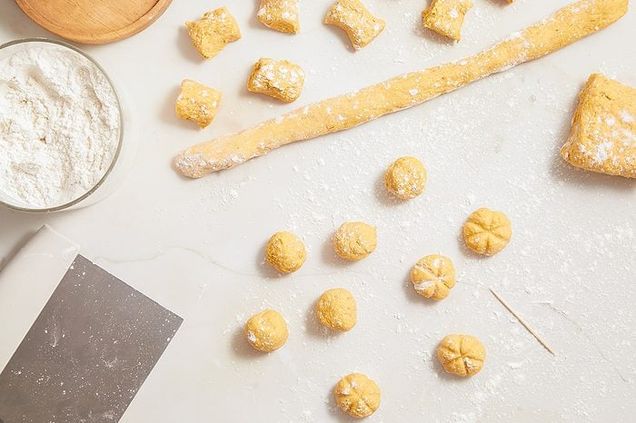 Overhead horizontal image of yellow gnocchi dough being prepared on a white, flour-dusted surface. A long roll of dough, cut dough pieces, and small shaped dough balls (some plain, some with grooved tops) are visible. Also present are a glass bowl of flour, a bench scraper, and a toothpick.