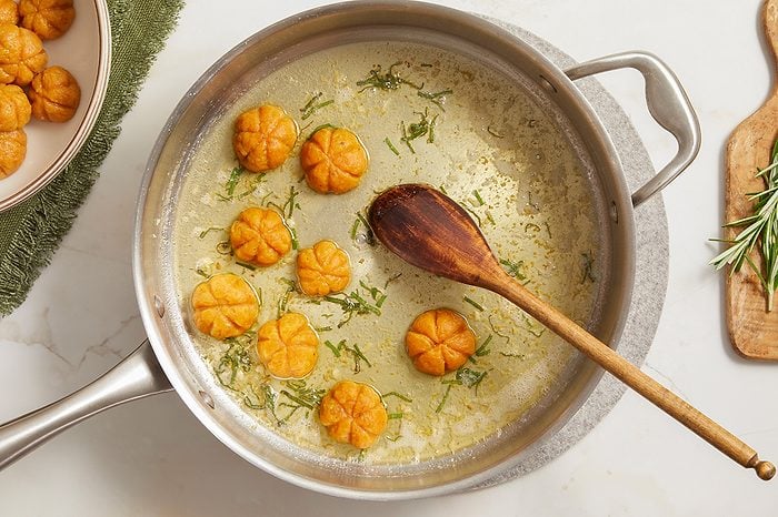 Overhead horizontal image of pumpkin-shaped gnocchi being cooked in a pan with a butter-herb sauce. Several of the small, orange, decorative gnocchi are in the pan alongside a wooden spoon, with more visible in a bowl to the left. The sauce contains chopped green herbs and garlic, and fresh rosemary sprigs are on a cutting board nearby.
