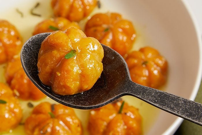 Eye Level horizontal image of a dark spoon holding one glossy, pumpkin-shaped food item over a white bowl. The bright orange food items are coated in a shimmering glaze and garnished with small green herbs, floating in a pale yellow liquid.