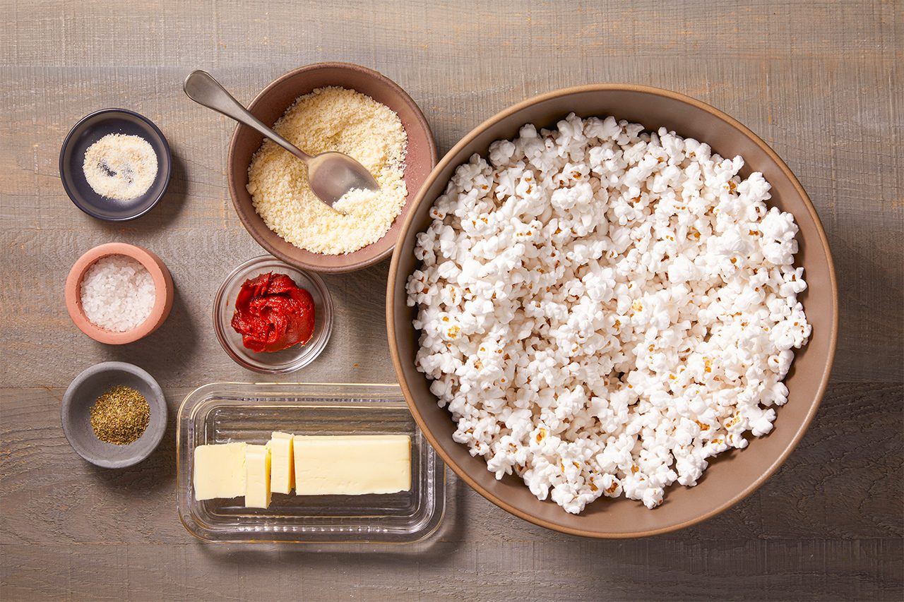A bowl of popcorn sits beside small dishes of seasonings, including grated cheese, red powder, salt, green herbs, garlic powder, and a few slices of butter, all on a wooden surface.