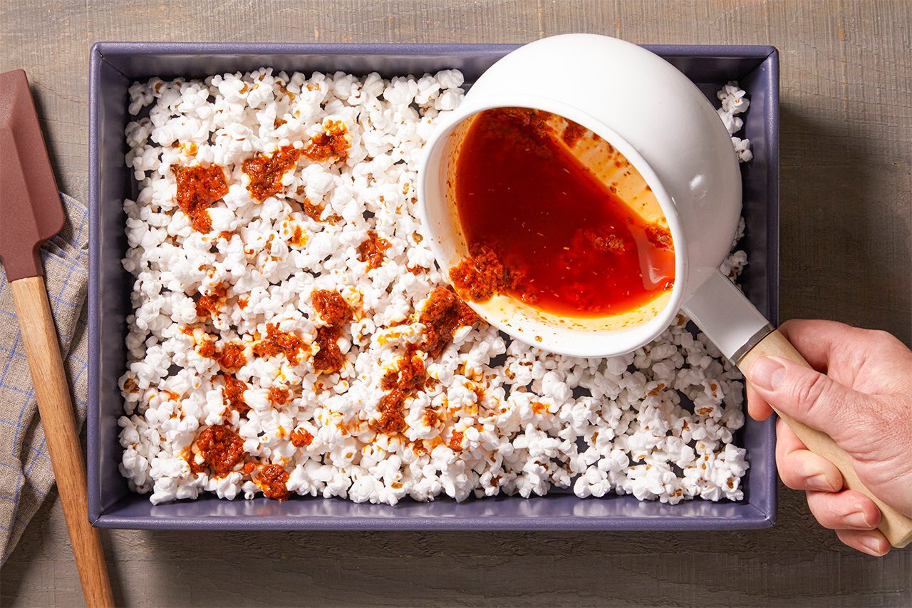 A hand pours red tomato sauce from a white saucepan over a rectangular dish filled with popped sorghum or small puffed grains. A spatula and towel are nearby.