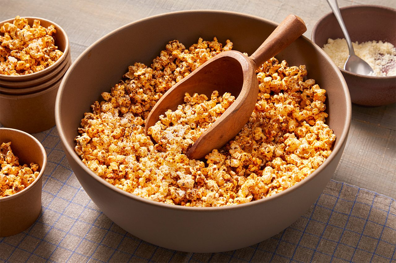 A large bowl of seasoned popcorn with a wooden scoop, surrounded by smaller empty bowls and a spoon in a small dish of grated cheese on a plaid tablecloth.